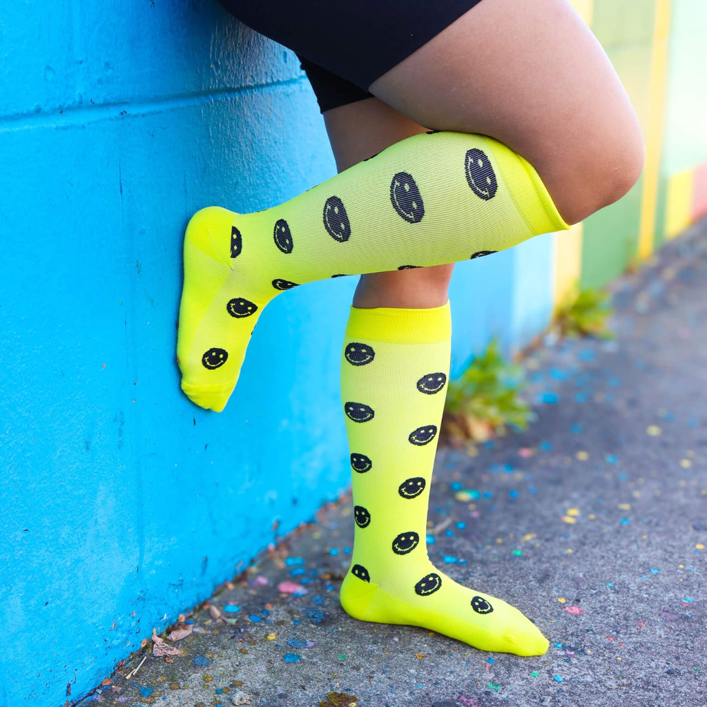 Woman wearing yellow compression socks with black smiley faces with foot against wall
